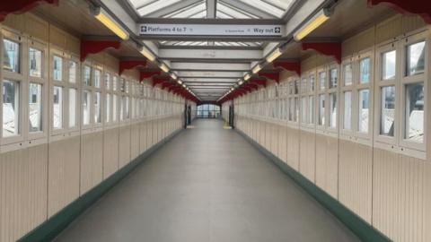 A view looking down a railway station footbridge length ways with windows either side. The walls are cream with a green skirting board and a grey floor.