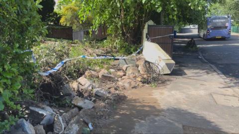 A front garden is taped off. There is rubble and ripped up hedges from a recent crash. A bus can be seen in the distance