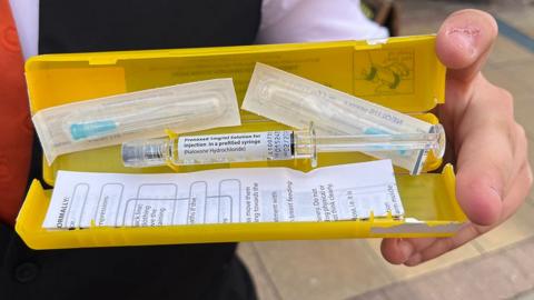 Person holding an anti-overdose kit. It is a yellow plastic box with a Naloxone filled syringe and two needles