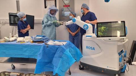 Four members of the operating theatre team prepare the robot ahead of surgery. The medics are wearing blue gowns and headwear. The robot has a white rectangular main body and an arm.