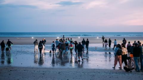 A large group of people walk along a beach towards the sea at sunrise or sunset. Their backs are to the camera.