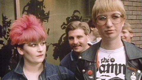 Two young women dressed as punks. One has pink hair and the other a blonde bowl cut and large glasses.