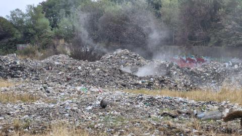 An image of a large pile of industrial waste in a field. The waste has small plumes of smoke coming from it, having recently been extinguished by fire crews.