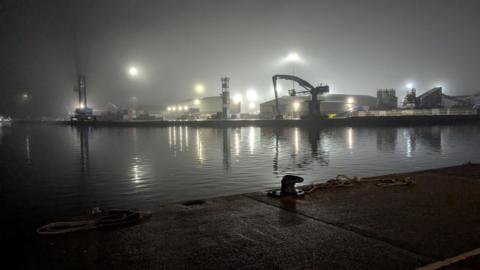 Poole quay at night in the mist. There are large floodlights over cranes and the light is reflected in the water of the harbour. 