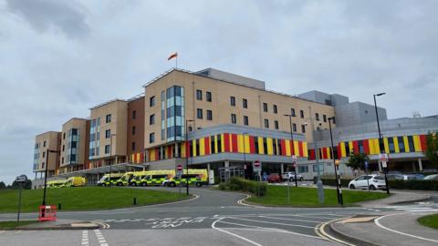 A hospital with ambulances parked outside it. The hospital building is several storeys high, and has red and yellow cladding on the lower part.
