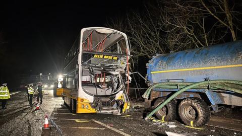 The double decker bus with both its front windows smashed and its back emergency door opened stands next to the end of blue tanker with surrounded by police tape and traffic cones with police officers in high visibility jackets standing by it