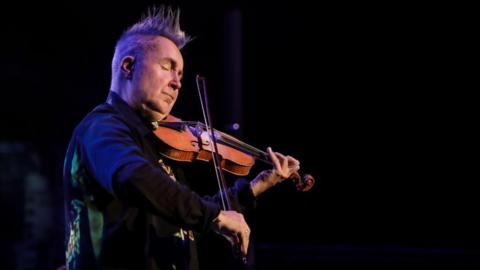 The image shows a man playing a violin with his eyes closed, wearing formal attire.