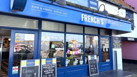 Front view of French’s Fish & Chips, a blue-painted seaside takeaway and restaurant with large windows, exterior lamps and entrance signs.