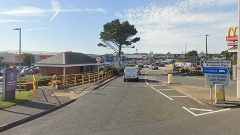 A Google Street image of a road leading towards a large car park. There is a sign on the right that says "Ravenside Retail & Park".