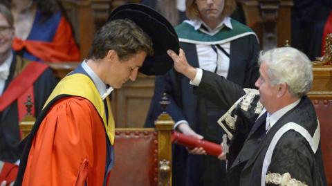 A person in a red academic gown bows during a university degree ceremony as an official in ceremonial robes lowers a black academic cap over the person's head, while attendees and faculty members look on in a wood-panelled hall.