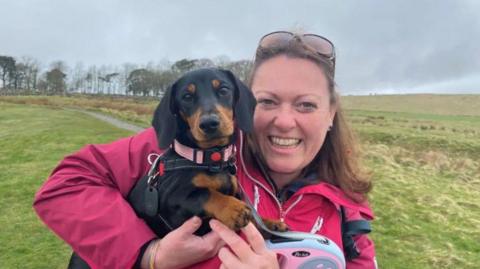 Kate Harrison is a young woman holding dachshund Bobbi in her arms. She is wearing a pink anorak and behind her is a long stretch of hillside with a path and a row of trees at the top 