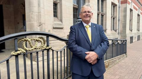 A man with long white grey hair in a dark navy suit, white shirt and yellow tie. He wears glasses and is standing outside a large light stone building with black metal fencing with good rope-type decorations.