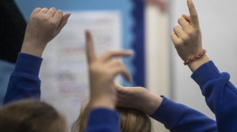 The hands of a number of children are in the air. They are wearing blue clothing and part of the heads of two children are pictured from behind.