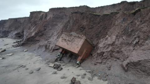 Still shot of the bunker at foot of the cliff. It is leaning into the collapsed cliff face and is surrounded by rock and sand.