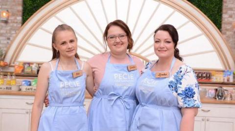 Three women stand side by side in a bright kitchen set, each wearing a light blue apron with a name tag. Behind them are white cabinets, baking equipment, and a decorative arch with a sunburst design.