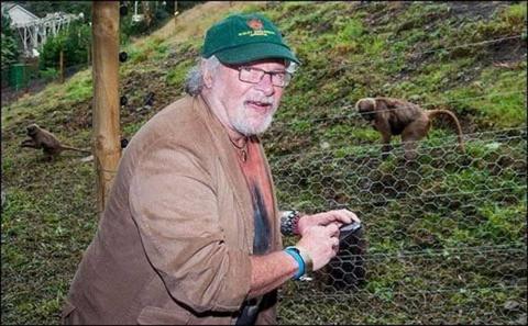 A man with grey hair and glasses with a green baseball cap and brown jacket. He is standing by an enclosure bounded by chicken wire, with a small primate on the grass behind. 