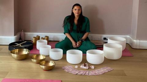 A woman in a green dress sits cross legged in front of pink yoga mats, white bowls and gold bowls before a wellness class. Two large pot plants are either side of the woman, who has long straight black hair.