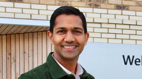 Anurag Aggarwal wearing a green jacket over a pink shirt. He has short black hair and is smiling in front of the hospice's cream brick building.