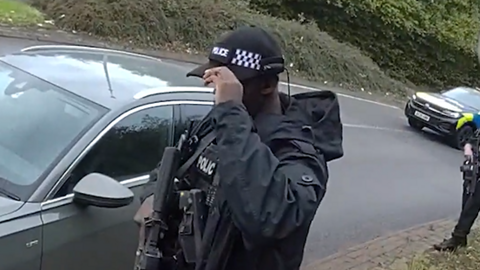 A police officer in black uniform, wearing a cap and face mask and carrying a large black gun in front of a silver car