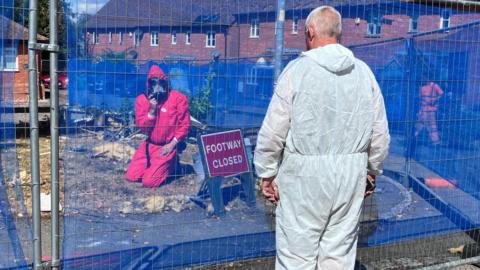 People dressed in white and pink hazmat suits are investigating the remains of an exploded bungalow with a blue fence surrounding