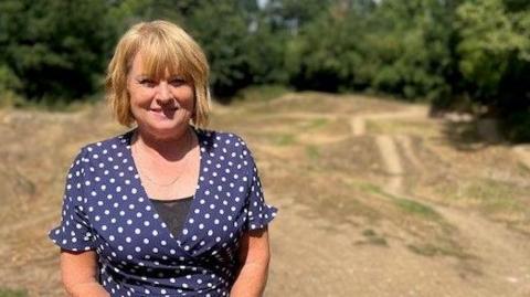 A woman with blonde hair wearing a blue polka-dot dress standing in front of a dirt bike track with mounds and trails. Green trees stand in the background.