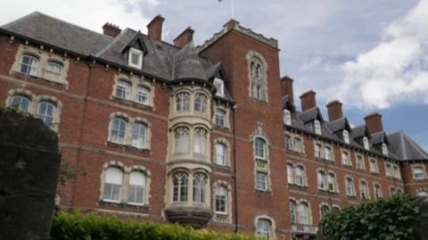 The image shows a large, heritage brick building with perhaps gothic features around windows and a tall central column in the centre of the school.