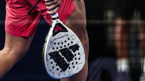 A padel raquet being carried by a player walking across a court, dressed in red, during the Paris Major Premier Padel tournament.