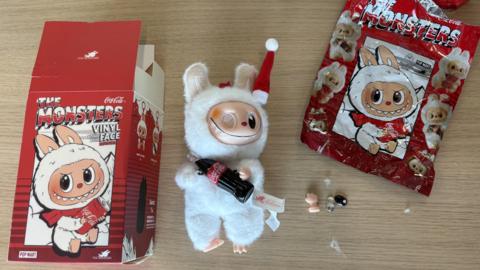 A fake Labubu doll rests on a wooden surface. It has a white plush body and plastic face. It is missing one eye. It holds a small Coca Cola bottle. One of its hands which has fallen off and its missing eye rest next to it along with a packaging box and plastic packaging bag.