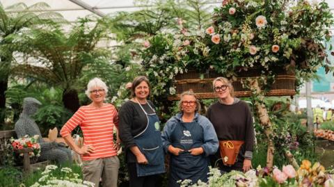 Four women standing in a line and smiling. Three are wearing glassed and two have blue aprons on. They are stood in front of a funeral flowers display which has flowers bursting out of an elevated coffin next to the a sculpture of a man sat on a bench.