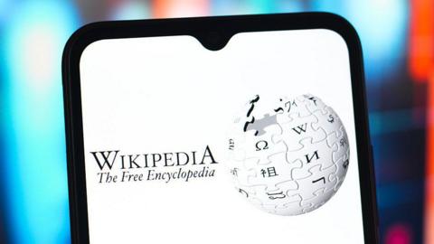A stock image showing a screenshot of Wikipedia's logo - a globe constructed out of jigsaw piece like elements bearing letters in different global alphabets -  on a mobile phone. The text in the image reads "Wikipedia The Free Encyclopedia"