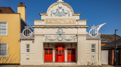 Edwardian cream-coloured building with red doors and Electric Palace in bas relief gold lettering above the door. The building is decorated with plasterwork of wreaths and vines, painted grey. 