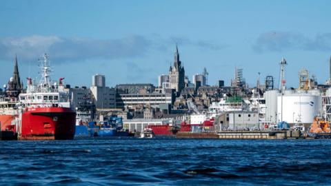 A view of Aberdeen waterfront and cityscape from close to the water just off shore. We can see two red-hulled ships, grey fuel storage tanks and grey granite buildings behind. It is a sunny day and the sea is very blue.