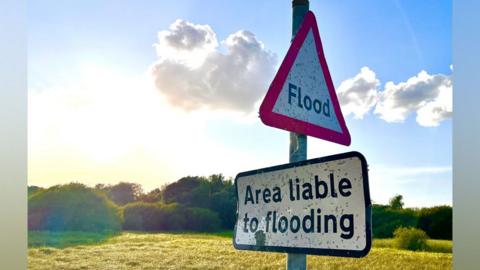 A bright sky over fields and hedges with two signs in the foreground. A triangular sign and a rectangular sign are stuck to a post. One reads "flood", the other says "area liable to flooding".