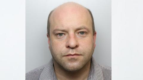 A police mugshot of Benjamin Baxter. He is balding, with short brown hair around the sides . He has stubble and wears an impassive expression.