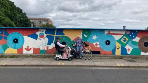 Two people on the pavement on Mill Road railway bridge in Cambridge, with a colourful mural on the bridge's side barrier