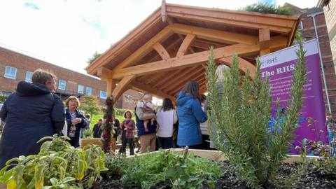Photo taken from inside one of the planters showing a range of shrubs in soil with people chatting at the garden's opening in the background.