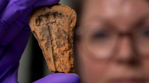 A woman, who is wearing glasses and her face blurred because she is out of focus, holds up the orange ceramic mould in a hand protected by purple plastic gloves.