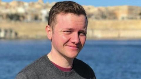 A man wearing a grey jumper and red top. He has short brown hair. He is standing in front of the sea, which is a bright blue, and a town in the background. The sky is blue. The background behind him is blurred. 