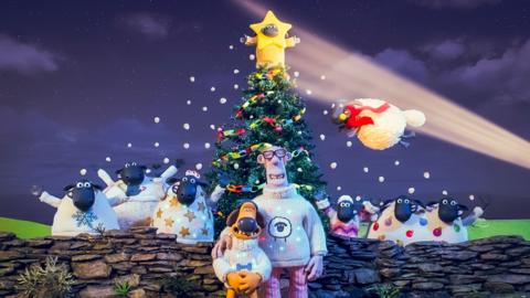 Shaun and the sheep behind a wall with decorated Christmas tree. The farmer is hugging bitzer and Timmy is ontop of the tree in a star costume