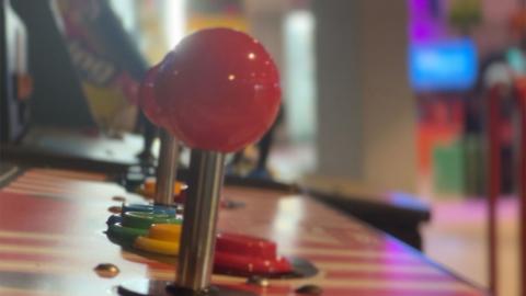 A close-up shot of the controls on an arcade games machine. A joystick with a red spherical handle is slightly blurred in the foreground - just in front of a row of red, yellow, green and blue push buttons and another joystick in the background.