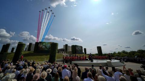 A crowd gathered by a large memorial in the National Memorial Arboretum. A military band can be seen next to a platform in the middle. The Red Arrows are rising up from behind the memorial in the background, emanating red, white and blue smoke trails.