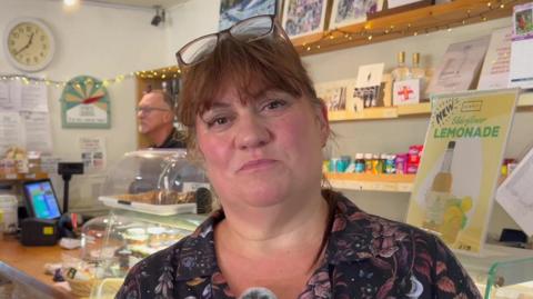 Manager Marie Humphries stood with glasses on her head in front of the shop's counter. There is a sign for elderflower lemonade behind her.