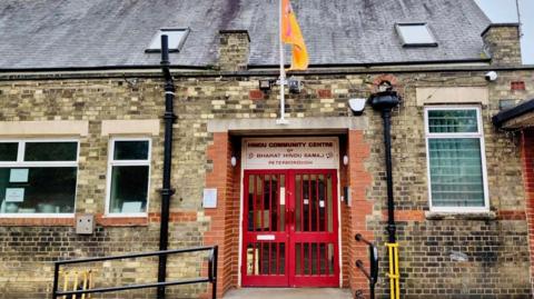 Exterior of the temple building with a red door. Bharat Hindu Samaj is written in white at the top of the door entrance and there is an orange flag.