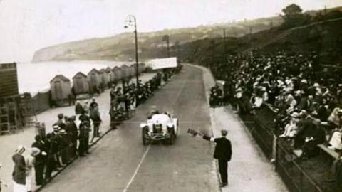 A white 1910s car races towards a marshall waving a flag at the finish line on Colwyn Bay prom. Bathing huts can be seen on the left, with a single layer of fans, whilst on the right is a railway embankment where spectators are massed seven or eight rows deep. 