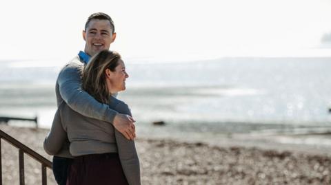 Peter Murphy has short black hair and is wearing a grey jumper. He has his arm around his wife, who has brown hair and is also wearing a grey jumper. They are standing by the beach in Shoeburyness.
