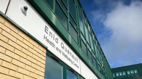 A photo looking up at the facade of the Enid Quenault Health and Wellbeing Centre. It shows the name of building on a white board, atop a brick wall. Above the name sign is dark green clad windows. The sky is blue with white clouds. 