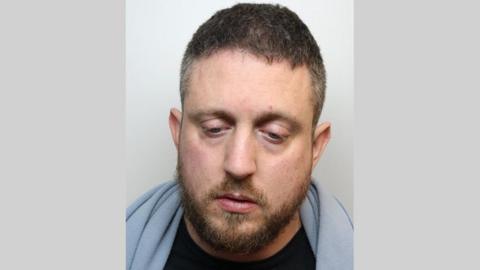 Custody image of Michael Elmer. he has a blue blanket around his neck and is looking down. He hs brown hair and a short, brown beard. 