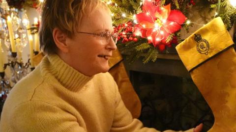 Andrea Middlemiss has short blonde hair and is wearing glasses and a yellow jumper. She is holding on the stockings that is fixed to a fire place decorated for Christmas. The yellow satin damask material has been made into a stocking with black edging and a crest.
