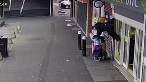 Lady hanging from the back of her coat outside a shop holding a trolley. She is horizontal and about four feet off the ground