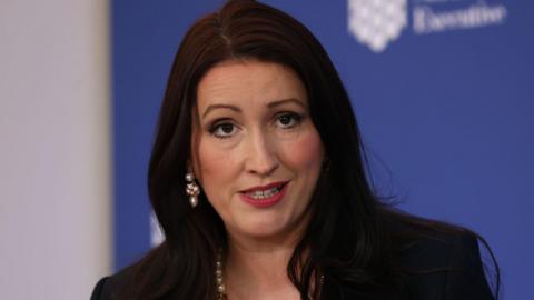 Northern Ireland Deputy First Minister Emma-Little Pengelly is speaking during a press conference. We can see the tops of her shoulders and head. She has shoulder-length dark hair and is wearing a pearl necklace and pearl earrings. She is wearing a dark jacket. In the background is a blue board with the logo of the Northern Ireland Executive